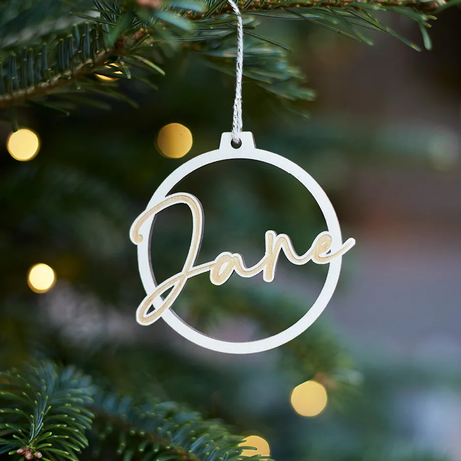 Een kerstbal uit hout gesneden met de naam Jane erin. Bal hangt in een kerstboom met lichtjes aan.